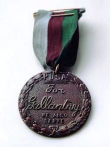 Bronze medal encircled in a laurel wreath and inscribed "PDSA For Gallantry We Also Serve" held from a ring suspender by a ribbon consisting of three equal vertical stripes of dark green, brown and pale blue