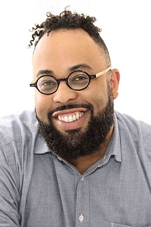 Headshot of Kevin Young. Young, with short hair and a beard, smiles at the camera; he wears a blue shirt and round glasses.