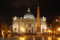 Behind a large monolithic obelisk, the facade of St. Peter's Basilica, lit by floodlights, rising majestically against the night sky