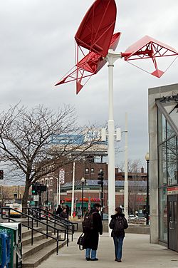 A slender white pole supporting three red, fan-like metal structures all on the same plane but twisted at different angles.