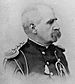 Profile of a balding white man with bushy, drooping mustache wearing an ornate military jacket with shoulder boards, shoulder cords, and a lanyard hanging from the chest.