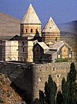 Stone church in the mountains with two massive towers topped by a conical roof.