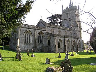 Gray stone building with arched windows and square tower. Foreground is grass with gravestones.