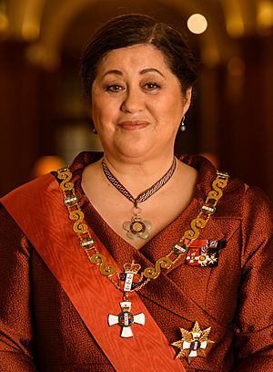 A smiling woman wearing a Māori feather cloak posing in front of a graphic of the New Zealand coat of arms