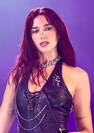 Headshot of Dua Lipa, a young, white woman with long, dark hair wearing a shimmery silver outfit and holding a microphone.