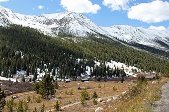 Several small wooden buildings in a valley with evergreens and snow-capped mountains in the distance