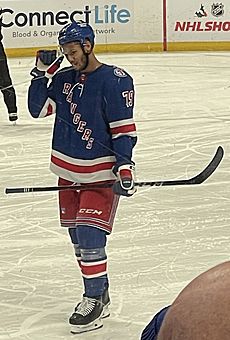 A young black man in a New York Rangers jersey holding a hockey stick in his left hand, looking down and to the side.