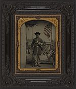 (Unidentified African American Union soldier with a rifle and revolver in front of painted backdrop showing weapons and American flag at Benton Barracks, Saint Louis, Missouri) (LOC) (5229147154)