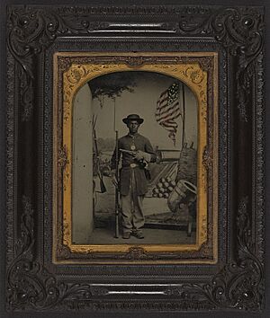 (Unidentified African American Union soldier with a rifle and revolver in front of painted backdrop showing weapons and American flag at Benton Barracks, Saint Louis, Missouri) (LOC) (5229147154)
