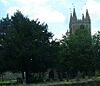 Top of ower with spirelets seen behind trees. In the foreground is grass and gravestones