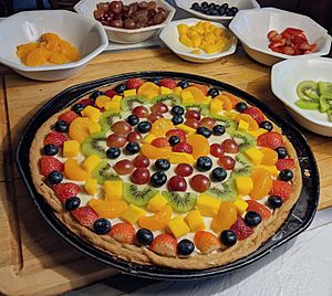 A fruit pizza on a kitchen counter and cuttong board, the fruit pizza surrounded by bowls of fruit peices that were used as toppings for the pizza.