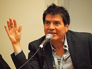 A middle-aged Chicano man with tousled hair speaks into a microphone. He has his right elbow resting on the table in front of him, his right palm facing out towards the audience.