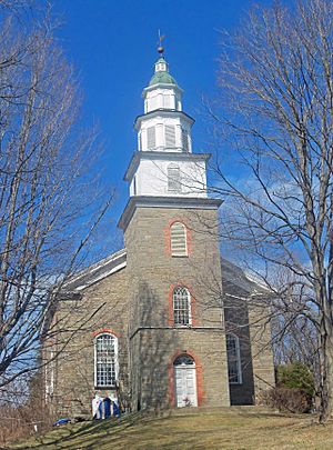 A church seen from the front on a clear day with some bare trees in front. All of it is stone except for the rounded window arches, trimmed in brick. Its steeple has wood painted white topped by a green cupola