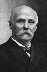 A black and white photograph of an elderly man with a white mustache in a suite and tie