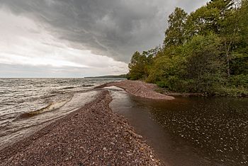 A stream of dark water flows from the right side of the images, away from the camera and some ways down a stony reddish beach, past a copse of green trees, before veering left across the beach and meeting a lake so wide that its far shores cannot be seen. The sky is cloudier on the right side, less so on left over the lake.