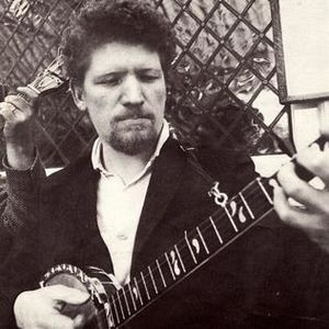 Black and white picture of a man (with focus on torso, not a full length picture). He has goatee (beard) and curly hair, and is holding a guitar He is looking at the guitar with concentration. He is wearing a shirt and coat.