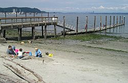 The public pier and waterfront at Manchester during a summer low tide. Bainbridge Island is in the background on the left, and Seattle is far away on the right.