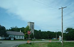 From the right turn lane on Taylor Road, the Bethel Bible Church at left, a rectangular grain elevator building in the center, and Illinois Route 8 sign on the right.