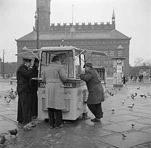 Mensen staan te eten bij een kiosk met op de achtergrond het raadhuis, Bestanddeelnr 252-8832