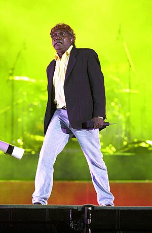A 44-year-old Aboriginal Australian man standing upon a stage, wearing light blue jeans, a black unbuttoned jacket, a yellow shirt and a headband. He holds a portable microphone in his left hand at his side and is staring ahead. Behind him is band equipment on a screen lit up in a green display.