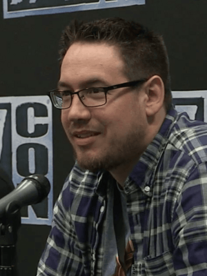 Brode wearing plaid and sitting behind a microphone against a background with BlizzCon logos