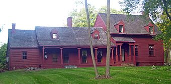 A red house in three sections. The middle has a porch, the right one has a second story. Two tree trunks are in front.