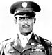 Head and shoulders of a black man, with a carefully trimmed mustache, standing erect and staring directly into the camera. He is wearing a peaked cap and a jacket with three rows of ribbon bars and a pin on the left breast, pins on the lapels, and a braided cord over the left shoulder.