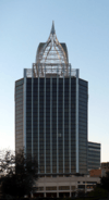 Ground-level view of a 30-story building with a square cross section and a dark glass facade; on the roof there is large, white, latticework structure that tapers into a spire.