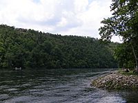 A wide river passes between a steep tree-covered hillside and a rocky shoreline, with houses built into the trees and fishermen in a canoe