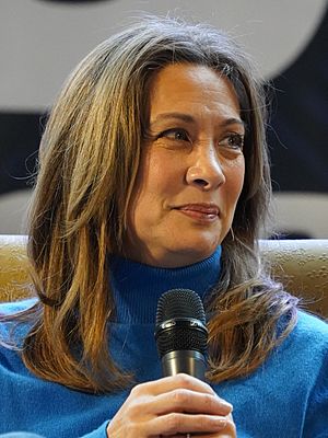 close-up of Diana Lee Inosanto wearing a royal blue turtleneck sweater, holding a microphone, smiling while looking left of camera