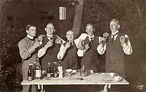 Fem män höjer sina snapsglas ute i en trädgård stående vid ett trädgårdsbord med kaffekoppar och flaskor - Nordiska Museet - NMA.0048890