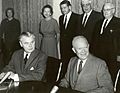 Diefenbaker and a smiling bald man in a suit sit at a table.  Two women and two men stand behind them.