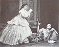 A man with a shaven head, wearing Asian dress, reclines on the floor and gestures at a woman in 19th century dress, who is writing, apparently at the man's dictation.