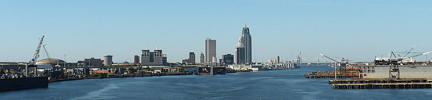 Panoramic view of a city's skyline; in the foreground, a large river and port facilities are present. In the distance, there is a city skyline with several skyscrapers of varying heights.