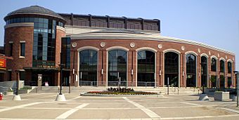 On the left is a two-storey red brick rotunda with a full-height five-pane wide dark window showing parts of the interior atrium, and at which base is the main entrance to the theatre. To the right is a red brick wall arcing away, with nine identical and equally spaced four-pane wide windows stretching from the ground to nearly the roof. It is fronted for its entire length by a stairway with about seven steps, leading to a piazza in the foreground.