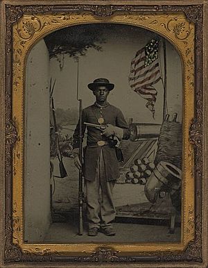 Unidentified African American Union soldier with a rifle and revolver in front of painted backdrop showing weapons and American flag at Benton Barracks, Saint Louis, Missouri LOC 5229147154 (cropped)