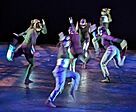Group of dancers in black and metallic colors in exaggerated running poses pointing in various directions
