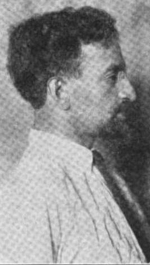 A photograph of a white man in profile; his hair is cut short, and his has a goatee; he is wearing a collared shirt and a necktie