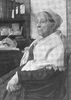 An older white woman, seated; she is wearing a head covering and a white shawl over a dark garment; one arm is resting on a desk with a quill pen in hand