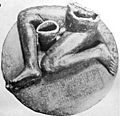 Black-and-white photograph of a statue consisting of an inscribed, round pedestal on top of which sits a seated nude male figure of which only the legs and lower torso are preserved.