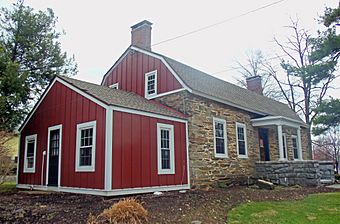 A small one-and-a-half-story stone building with a gambrel roof and one-story gabled wing on its left side photographed at an angle so that side is closer to the camera. Two chimneys rise from the roof; the wing and the roof apex on the side facing the camera are faced in vertical red boards.