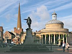 Statue of figure with outstretched arm. To the left a tall church spire and to the right a circular building with columns.