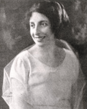 A smiling white woman with dark bobbed hair, wearing a white dress with a translucent shawl or wrap