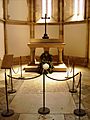 Photograph of a small chapel in which a roped-off inscribed stone slab is set into the floor and with a stone altar in the background on which are a wreath and a large metal cross