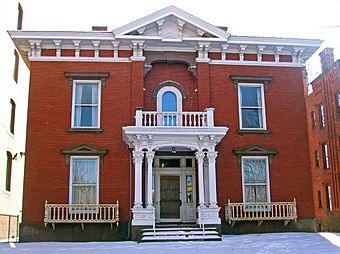 A brick house with white ornate wooden trim seen from the front. Its roof has a small pointed section in the middle, and the front door is boarded over. There is snow on the ground in front and the steps.