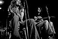 A black and white photograph of Robert Plant with a tambourine and Jimmy Page with an acoustic guitar seated and performing.
