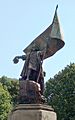 A photograph of a park in which stands a large statue depicting a bearded man standing on a rock who is dressed in a long coat and holding a hat in his right hand, while his left hand grasps a large banner