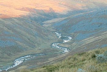 Ribbon of river in shadow reflecting the sky, bending back and forth through a valley between moorland hills of grey scree and brown and green vegetation