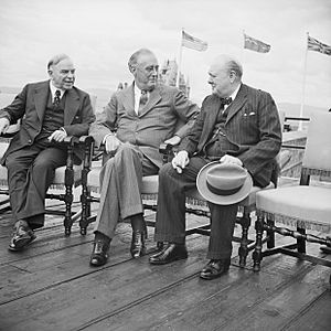 The three leaders sit on chairs on a wooden deck. Their national flags fly in the background.