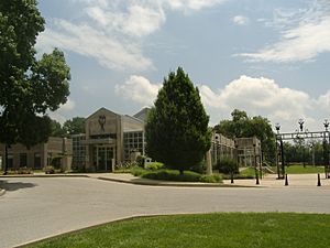 A rectangular conservatory building faced with cut limestone, viewed from an angle with the shorter side, which contains the entrance, to the left, and the longer side to the right. A large tree stands in front of the corner of the building. On the right side of the picture is the metal arched gateway to a plaza that is on the longer side of the building.
